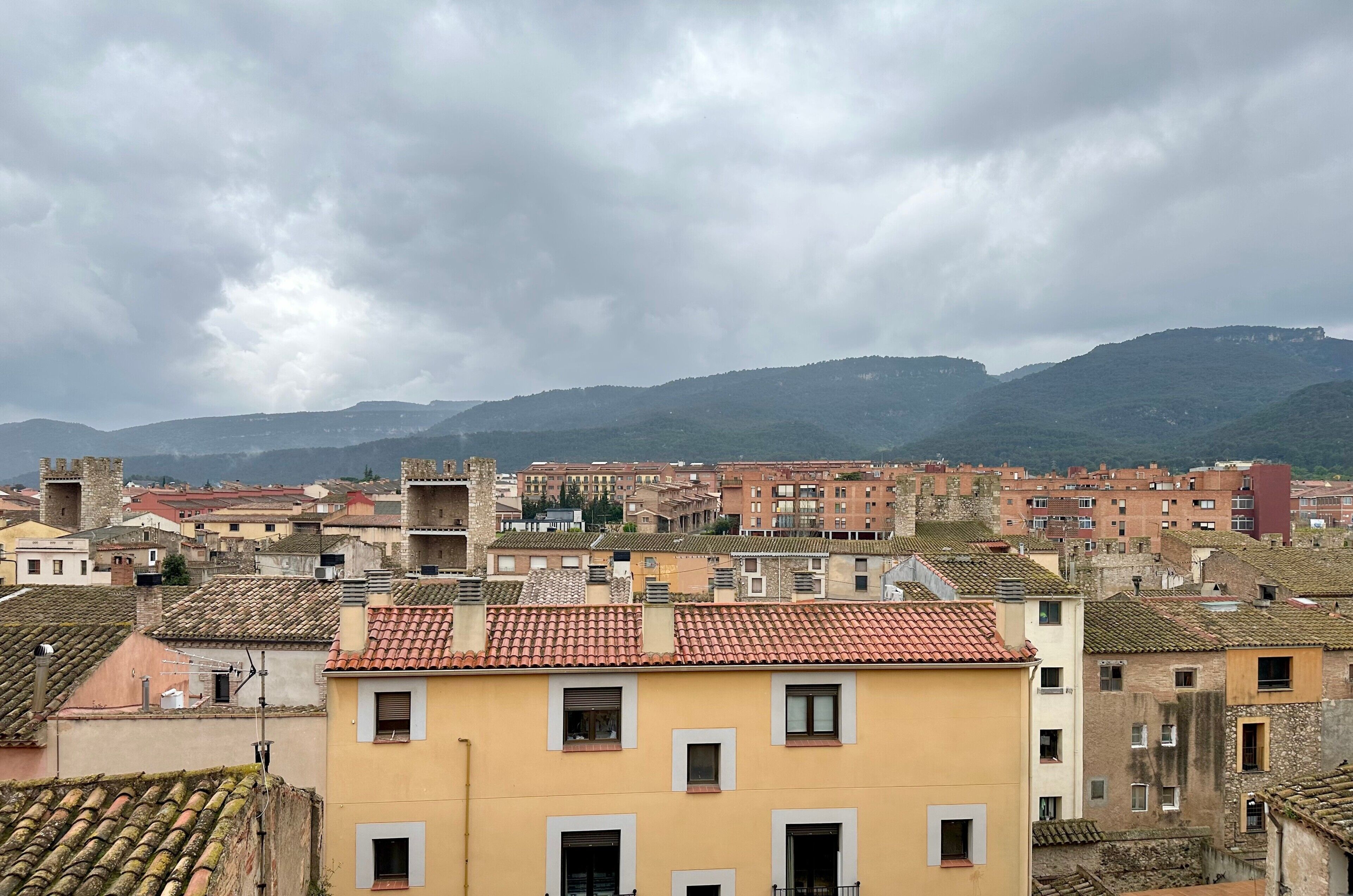 A cityscape with historic buildings, terracotta roofs, and a mountainous backdrop.