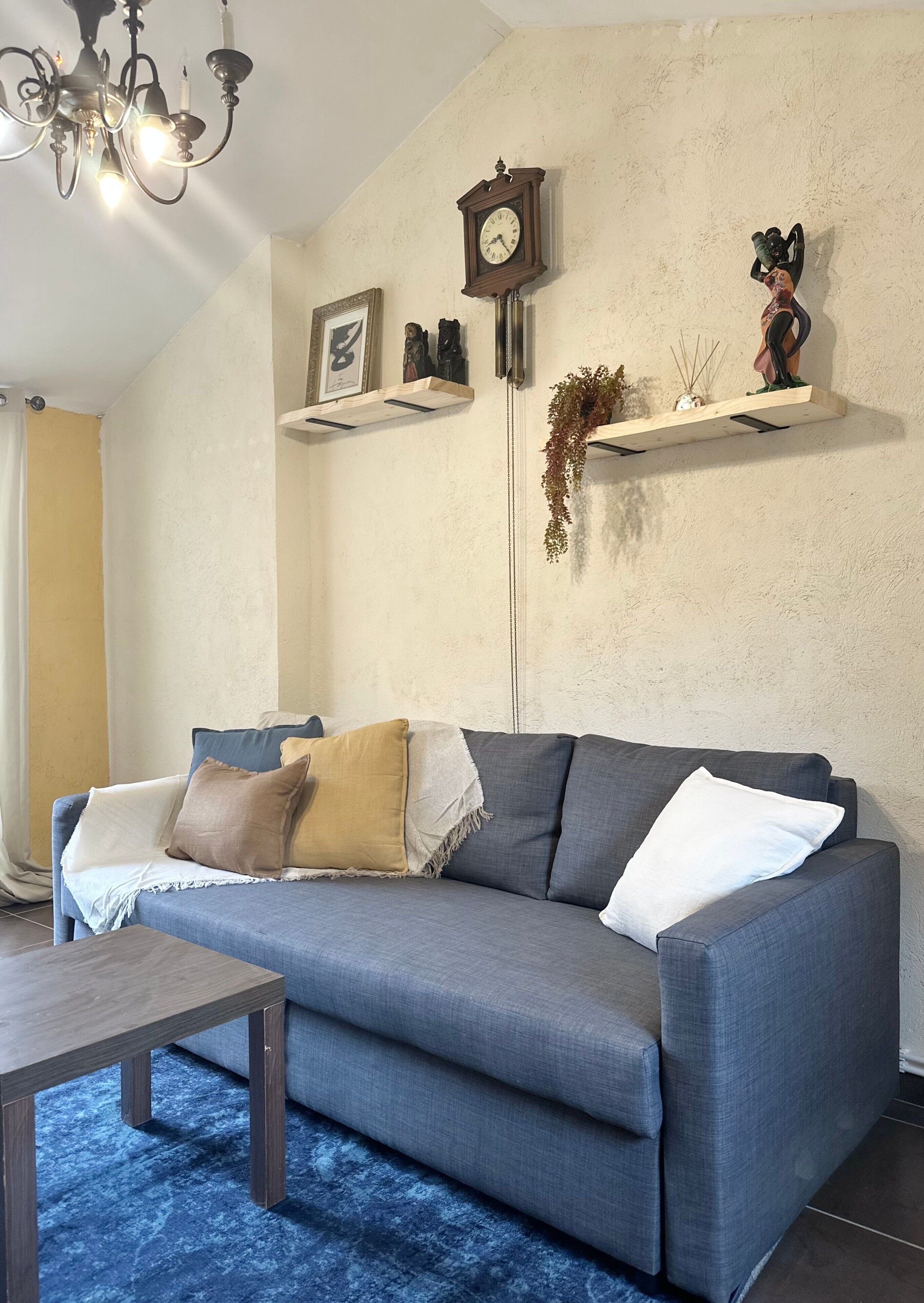 Een woonkamer met een grijze bank, een houten salontafel, een kroonluchter, wandplanken met decoratieve objecten, en een klok.
