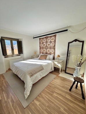 Une chambre à coucher avec un lit, une fenêtre, une table de chevet, un miroir et un vase avec des plantes.