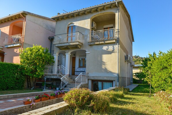 A two-story building with balconies, a front entrance, and a small garden area with potted plants.