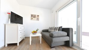 Ett modernt vardagsrum med en platt-TV, ett vitt skåp, en grå soffa, ett litet vitt bord med en fruktskål och ett konstverk på väggen.
