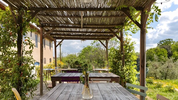 Une table en bois, un vase, une pergola et, en arrière-plan, une vue sur la verdure et un bâtiment.