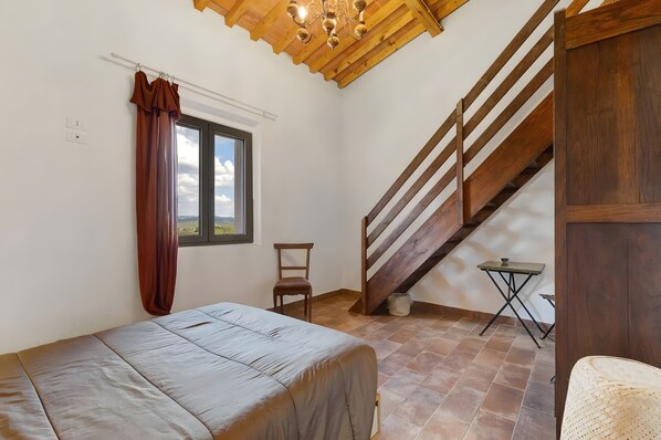 Un dormitorio con cama, una silla, una mesita, un ventanal con cortinas, una escalera de madera y vistas al exterior.