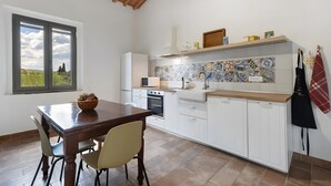 Une cuisine avec des meubles blancs, une table en bois, des chaises, une fenêtre donnant sur un beau paysage et un dosseret carrelé.
