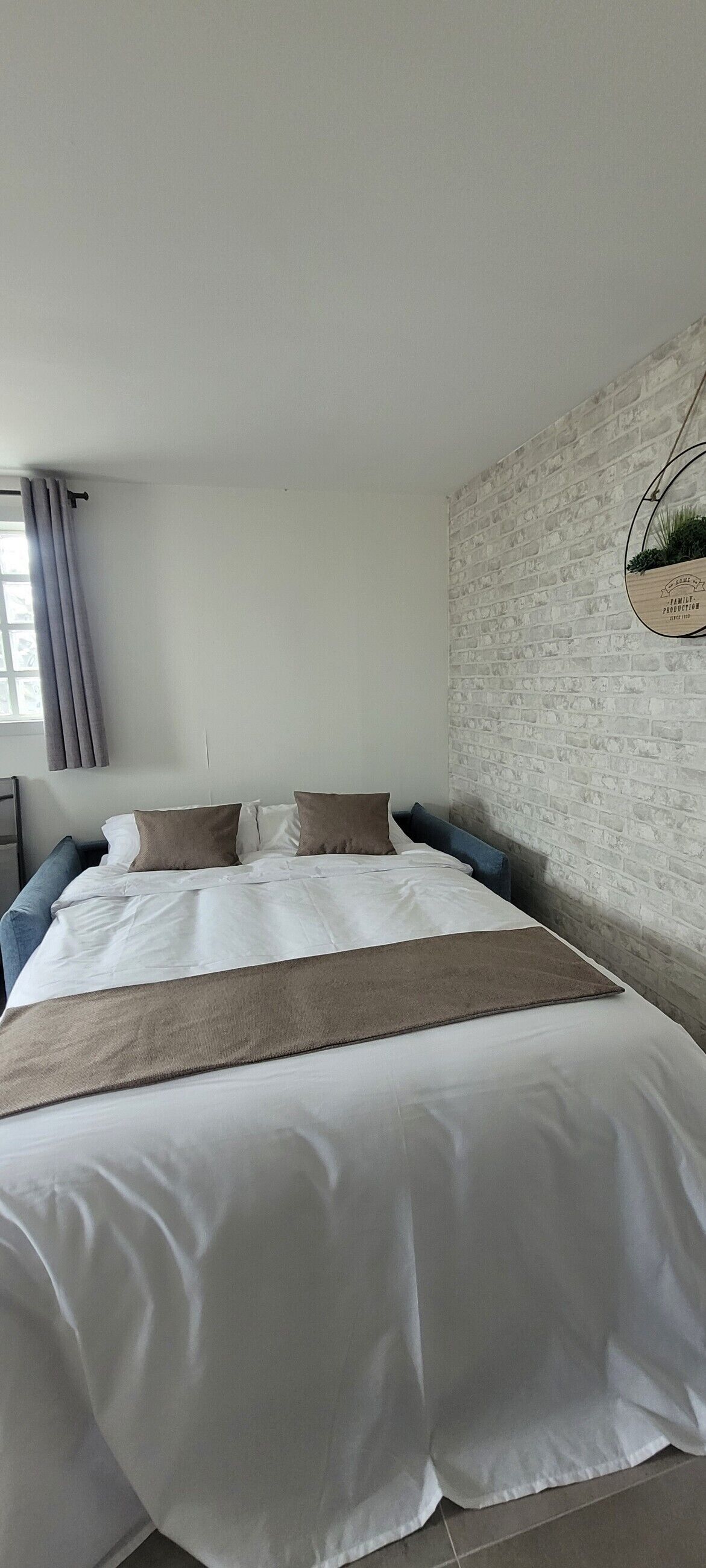 Un letto ben rifatto con lenzuola bianche e una coperta marrone, una tenda grigia, un muro con decorazione a mattoni e una pianta sospesa.