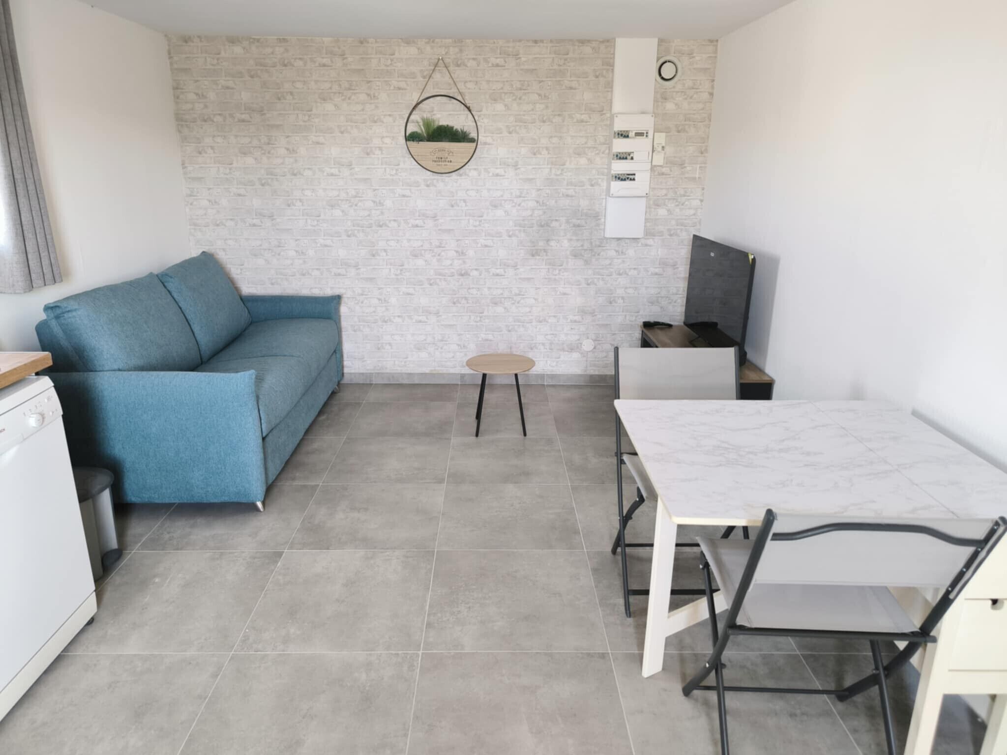 Un espace de vie compact comprenant un canapé bleu, une petite table ronde, une table à manger blanche avec des chaises, et un mur décoré d’un motif en briques sur lequel est accrochée une décoration circulaire.