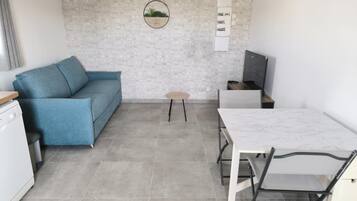 Un espace de vie compact comprenant un canapé bleu, une petite table ronde, une table à manger blanche avec des chaises, et un mur décoré d’un motif en briques sur lequel est accrochée une décoration circulaire.