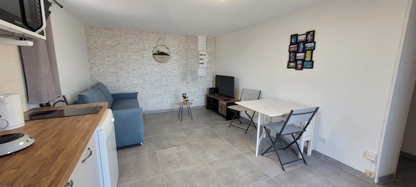 Un espace de vie compact comprenant un coin cuisine, un coin repas, un coin salon avec un canapé et une petite table, ainsi qu’un téléviseur.