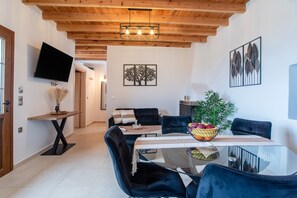A modern dining area with a glass table, dark upholstered chairs, a wooden ceiling, and a flat-screen TV mounted on the wall.
