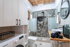 A modern bathroom with a washing machine, a glass-enclosed shower, a sink, and a wooden shelf with towels.