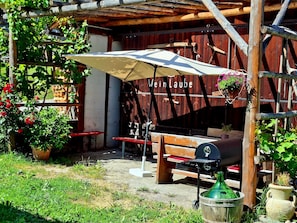 A rustic outdoor seating area with a wooden pergola, a barbecue grill, and a sign that reads 'Weinlaube.'