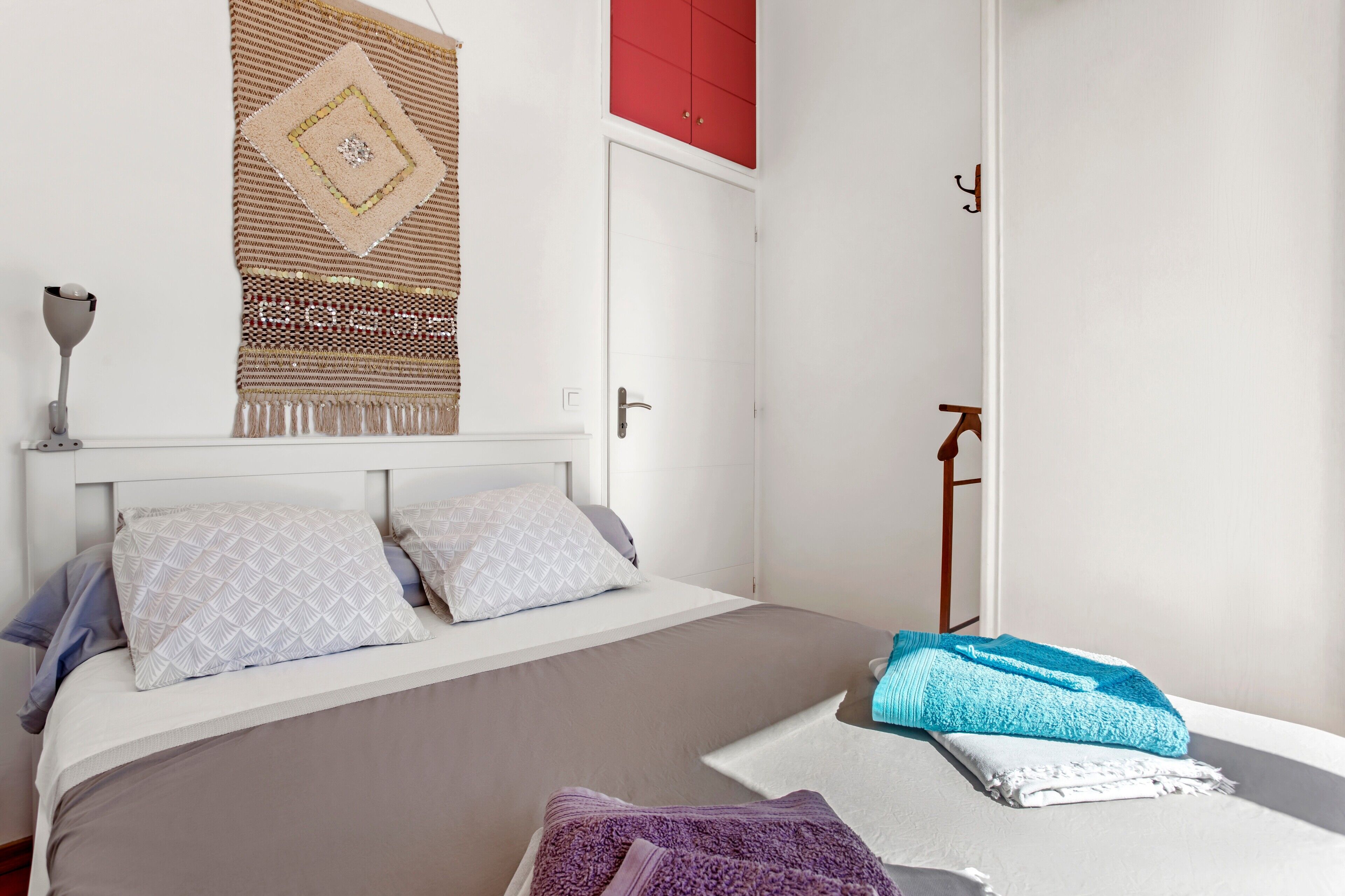 A neatly made bed with patterned pillows, a hanging tapestry, and a red wardrobe.