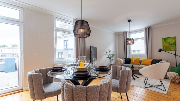 A modern living room with a round glass dining table, comfortable seating, and large windows offering a view of the city.