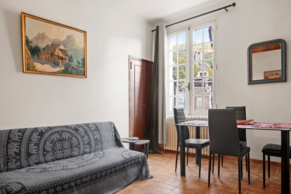 A room with a sofa, dining table, chairs, a painting, and a window with curtains.