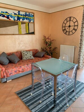 Una stanza con un divano, un tavolino in vetro, sedie trasparenti, una sveglia e un termosifone.
