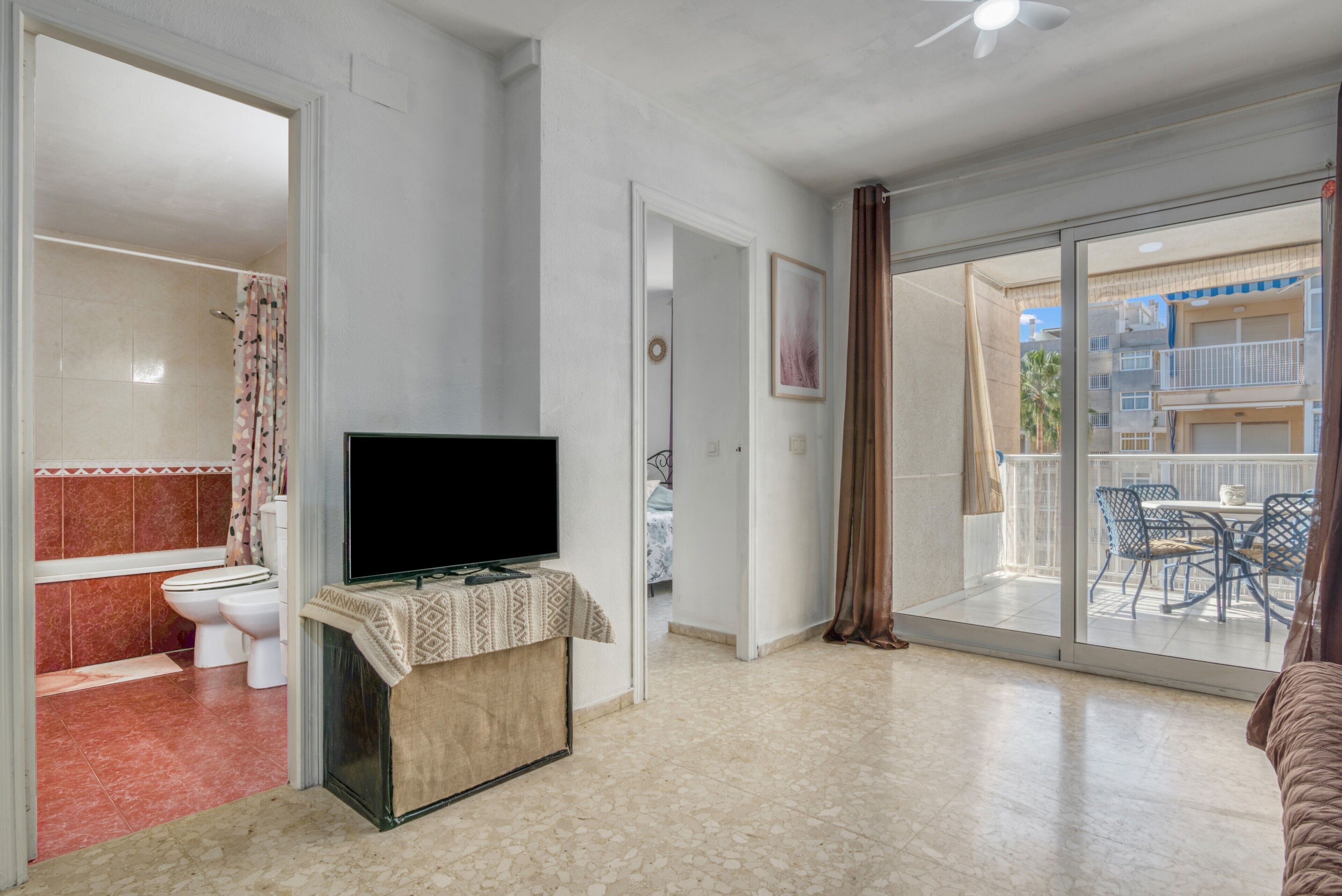 Ein Zimmer mit Fernseher auf einem Schrank, ein Badezimmer mit Dusche und Toilette sowie ein Balkon mit Gartenmöbeln.