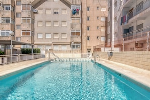 A swimming pool surrounded by a multi-story building with balconies, a covered area, and a tiled walkway.