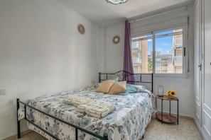 A bedroom with a bed, a window with a view of buildings, a small table with a vase and a candle, and a curtain.