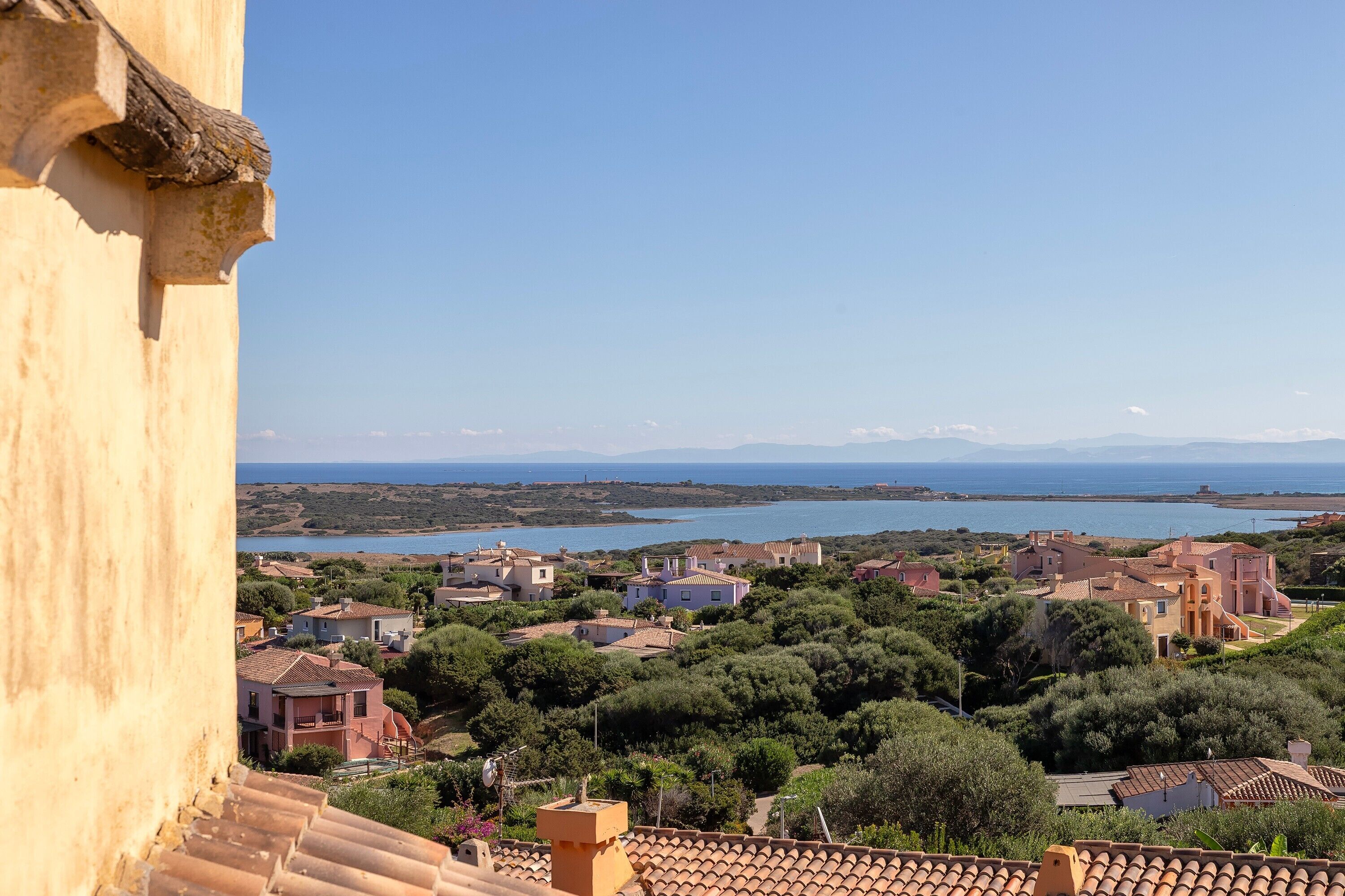 A coastal town with terracotta-roofed buildings, a clear blue sky, and a view of the sea.