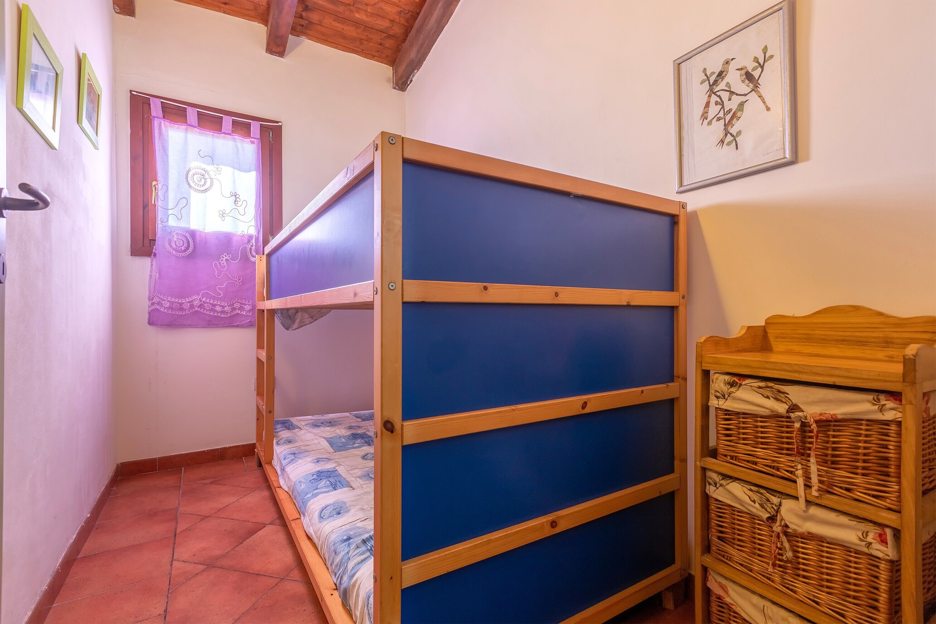 A room with a bunk bed, a window with a curtain, a framed picture on the wall, and a wooden chest of drawers.