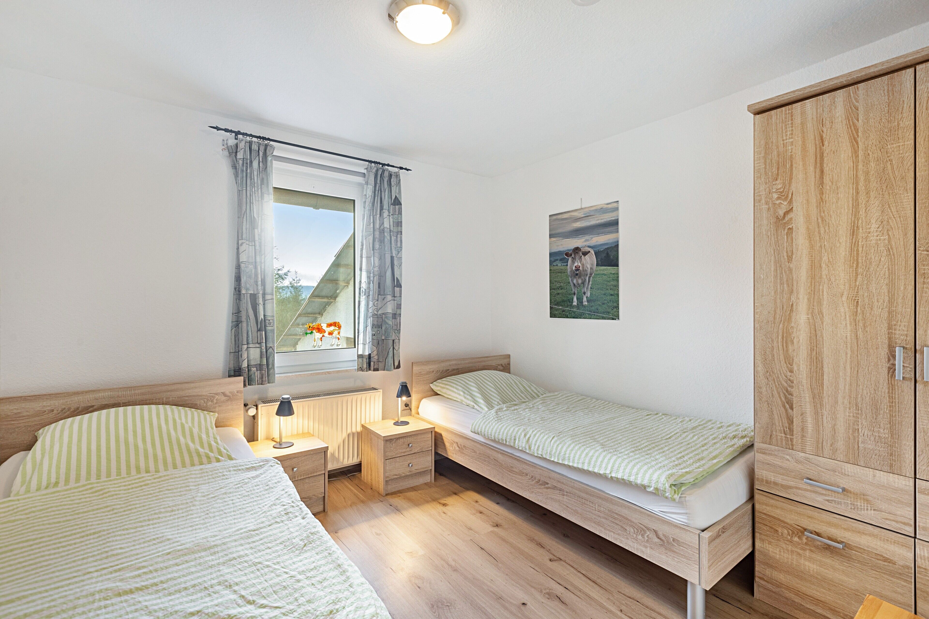 A bedroom with two single beds, a nightstand with a lamp, a window with curtains, a wooden wardrobe, and a painting of a cow on the wall.