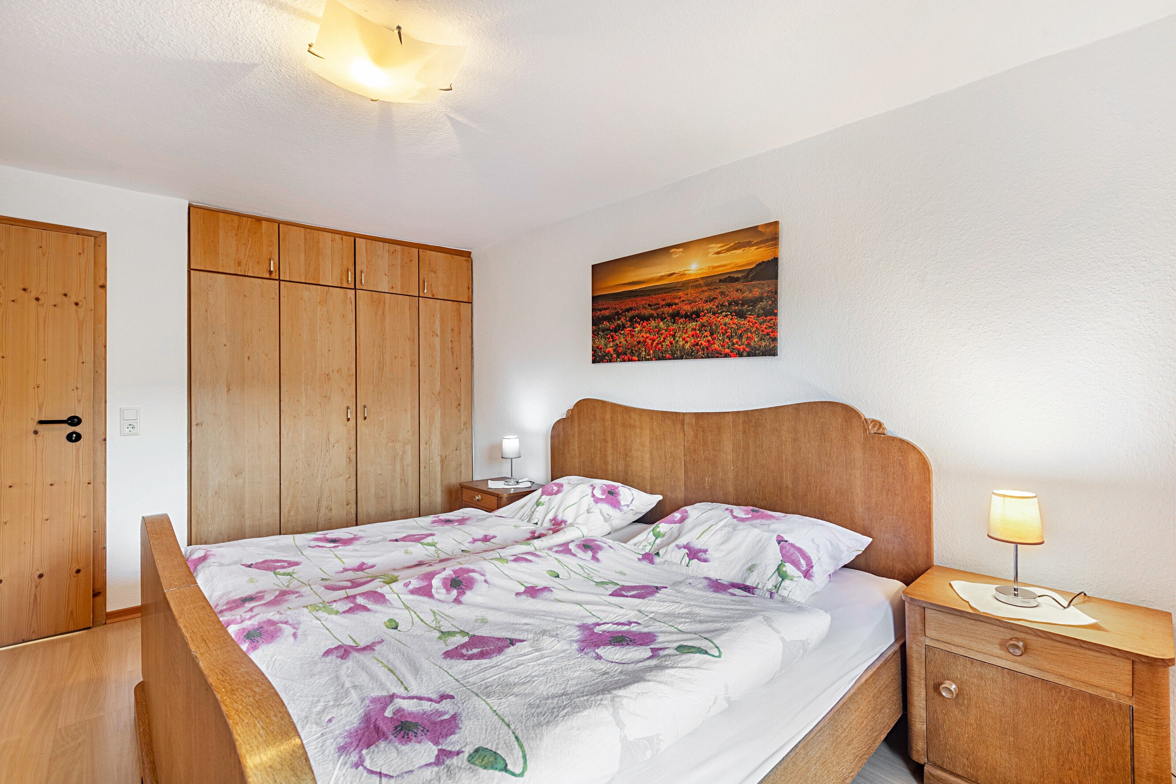 A bedroom with a wooden bed, a nightstand with a lamp, a wooden wardrobe, and a wall painting.