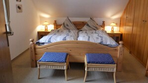 Un dormitorio con una cama de madera, dos mesitas de noche con lámparas, un armario de madera y dos sillas de mimbre.
