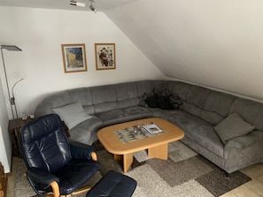 Un salon moderne avec un canapé d’angle gris, une table basse en bois, un fauteuil en cuir noir et un lampadaire. Deux tableaux encadrés sont accrochés au mur.