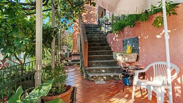 Eine Terrasse mit gefliestem Boden, einer Treppe, Kübelpflanzen, einem weißen Plastikstuhl und einem Grill.