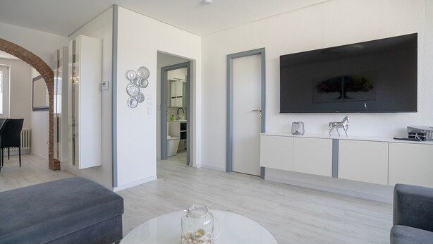 Una sala moderna con una mesa redonda blanca, sofás grises, un televisor de pantalla plana grande y una cocina al fondo.