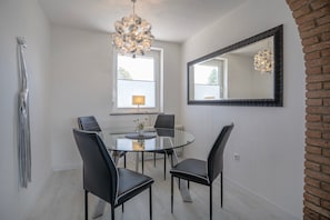 A dining area with a round glass table, four black chairs, a modern chandelier, a large mirror, and a window with blinds.