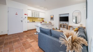 Una sala de estar moderna con un sofá, televisión y área de cocina con gabinetes blancos y salpicadero de azulejos.