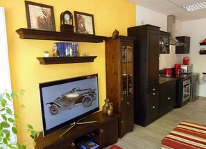 Una cocina con horno integrado, microondas y cafetera, un armario de madera con puertas de cristal, y un televisor mostrando un automóvil.