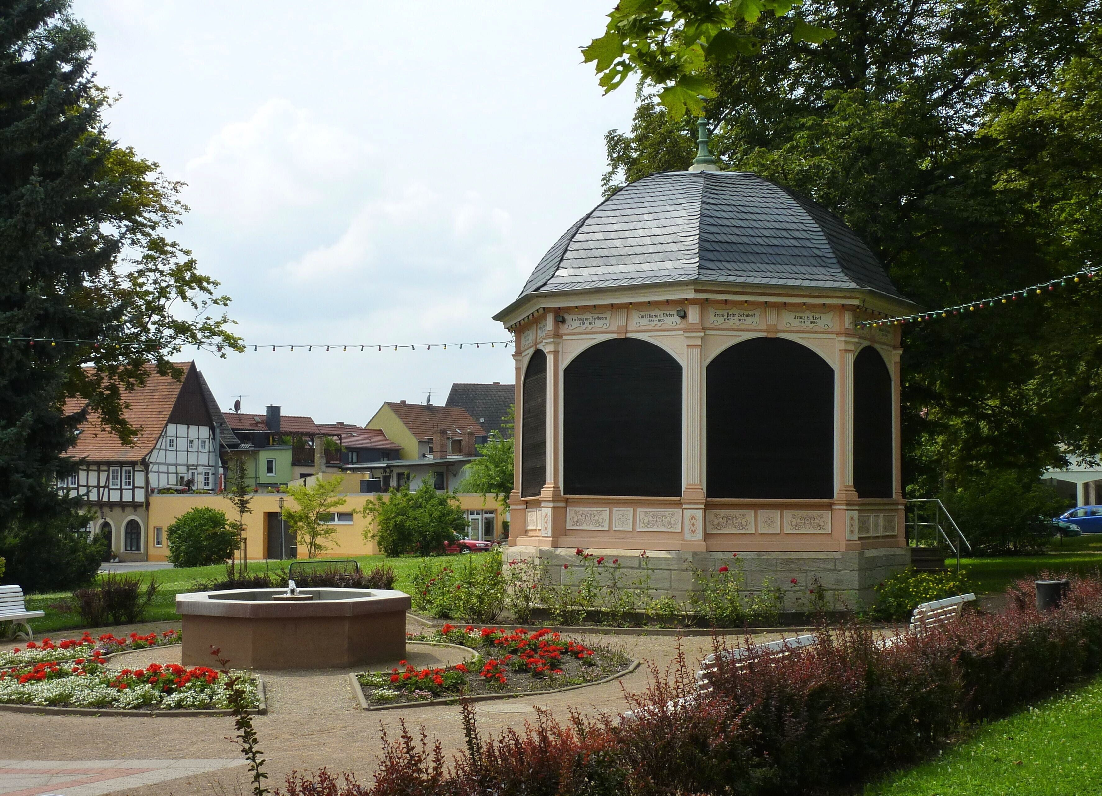 Ein Gazebo mit einem Ziegeldach, umgeben von einem Blumenbeet und Bänken, in einem Park mit Wohnhäusern im Hintergrund.