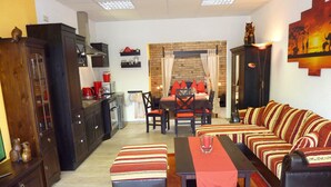 Un salon comprenant un canapé, un pouf et une table basse, un coin repas avec une table et des chaises, et une cuisine équipée de meubles et d’appareils électroménagers.