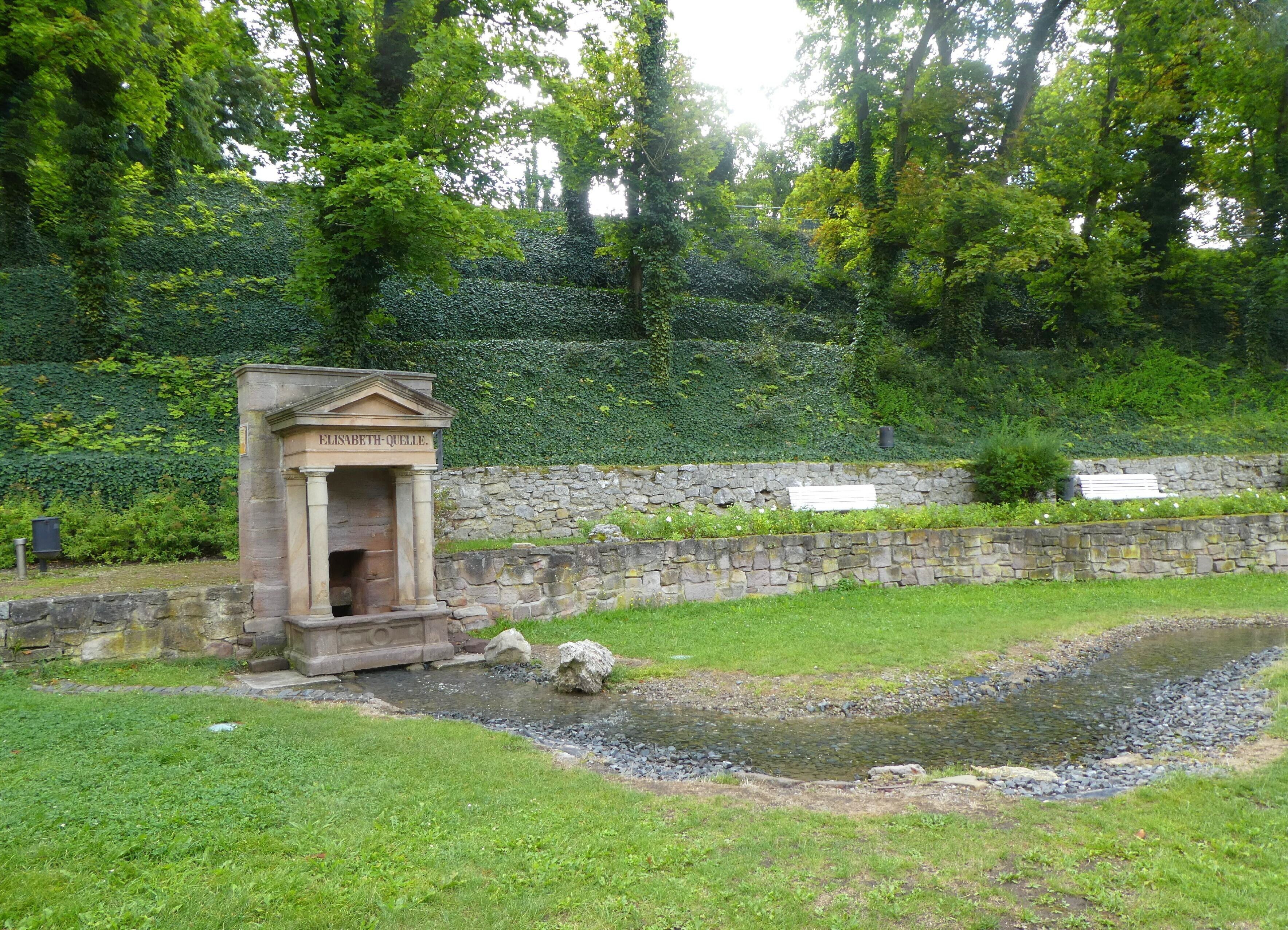 Une structure en pierre portant une plaque avec une inscription, un petit étang et un mur de pierre orné de verdure.