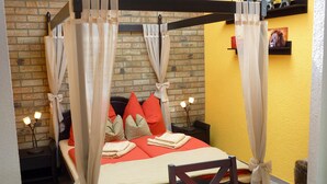 Un lit à baldaquin à quatre poteaux avec une literie rouge et beige, un mur en briques et un mur d’accent jaune.