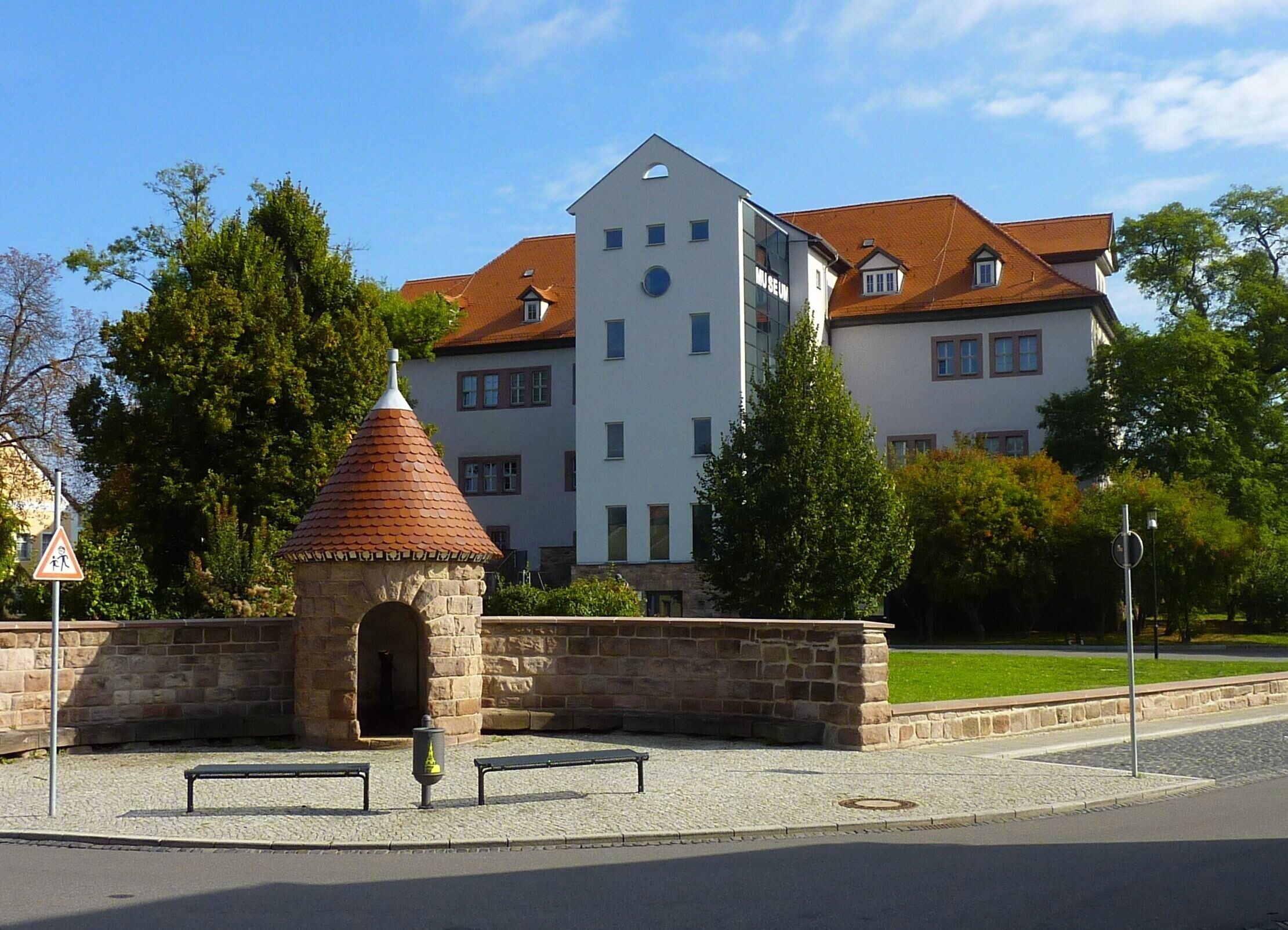 A stone structure with a conical roof, benches, and a building with a clock tower in the background.