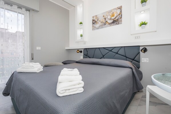 A modern bedroom with a grey bed, white towels, a grey headboard, and a round white table with a glass top.
