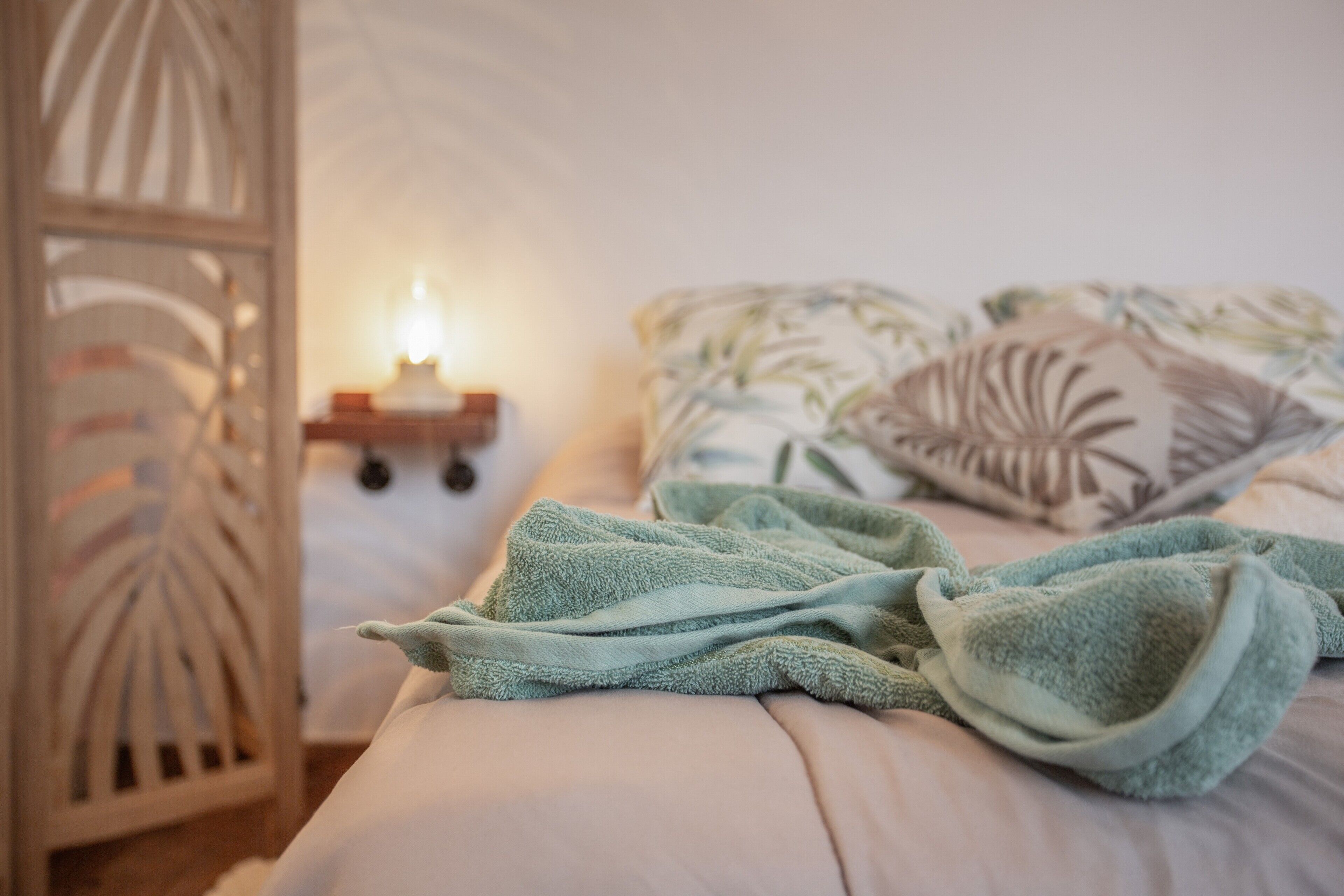 Una cama con una toalla verde, un cojín floral y una mesita de noche con una vela.