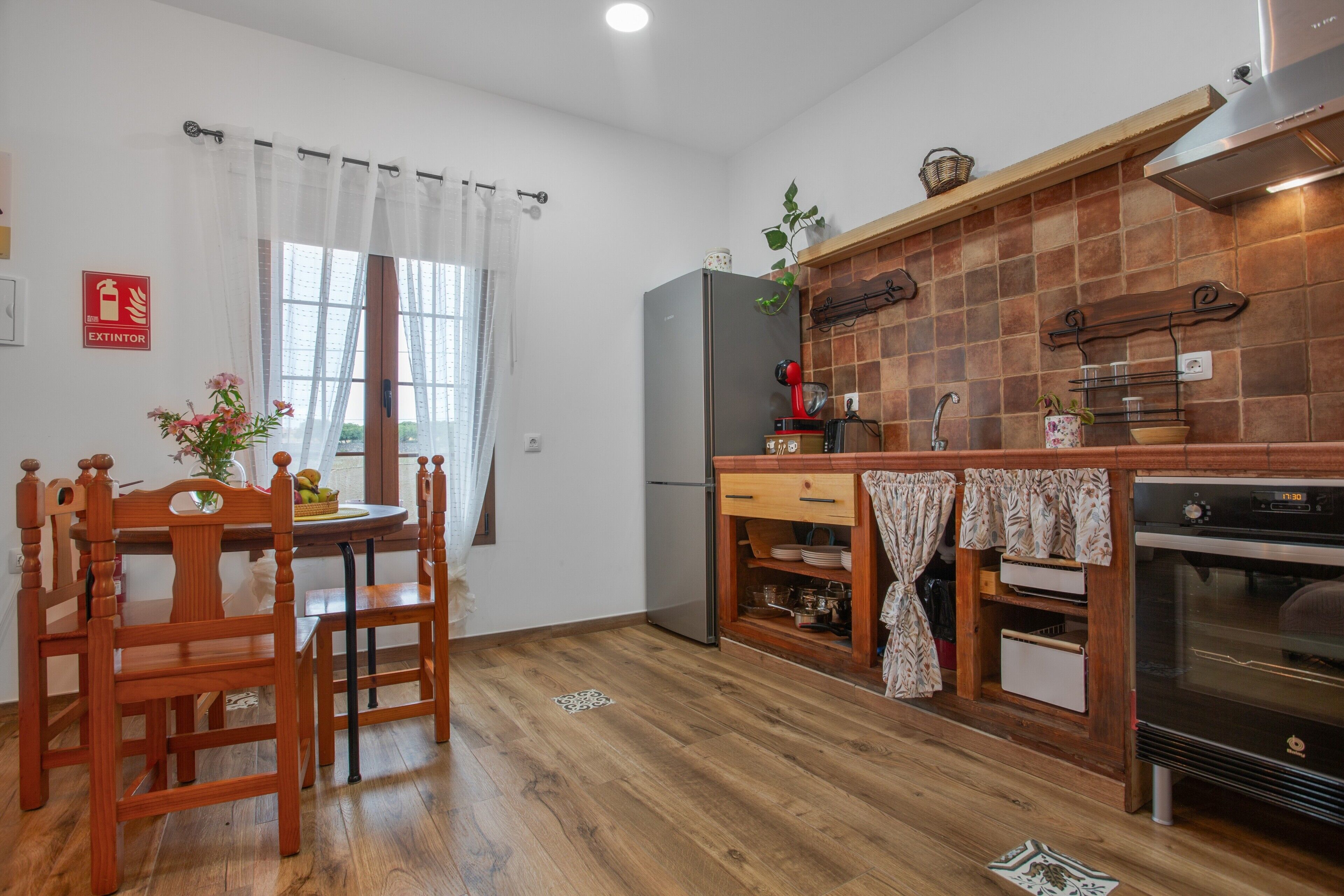 A kitchen with wooden flooring, a refrigerator, a stove, and a dining area with chairs and a table.