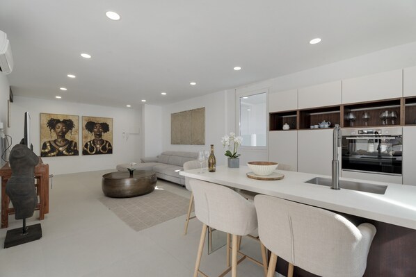 A modern kitchen with a white countertop, bar stools, a built-in oven, and a large window.