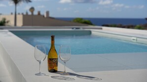 Escena junto a la piscina con una botella y dos vasos, con vista a un cielo azul claro y al océano.