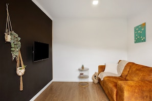 A living room with a brown sofa, a flat-screen TV, a small round table with a plant, and a wall-mounted plant arrangement.
