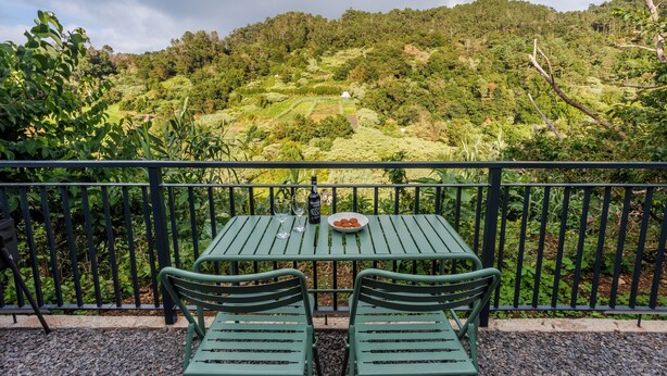 A green metal table with two chairs, a plate of food, and two wine glasses on a balcony with a view of a lush green hillside.