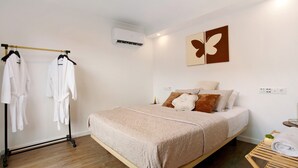 A bedroom with a bed, a wooden floor, a wall-mounted air conditioner, a towel rack with robes, and a wall art piece.