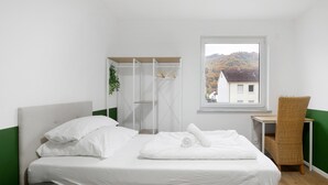 Uma cama bem arrumada com roupa de cama branca, uma mesa pequena com uma cadeira, uma janela com vista para edifícios e colinas, e uma parede verde.