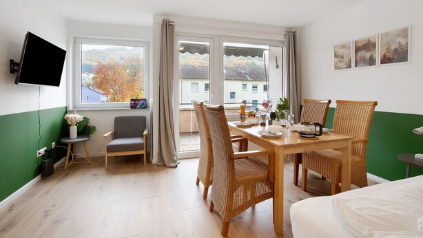 Quarto de hotel com mesa de jantar, cadeiras, televisão e janela com vista para edifícios e árvores.