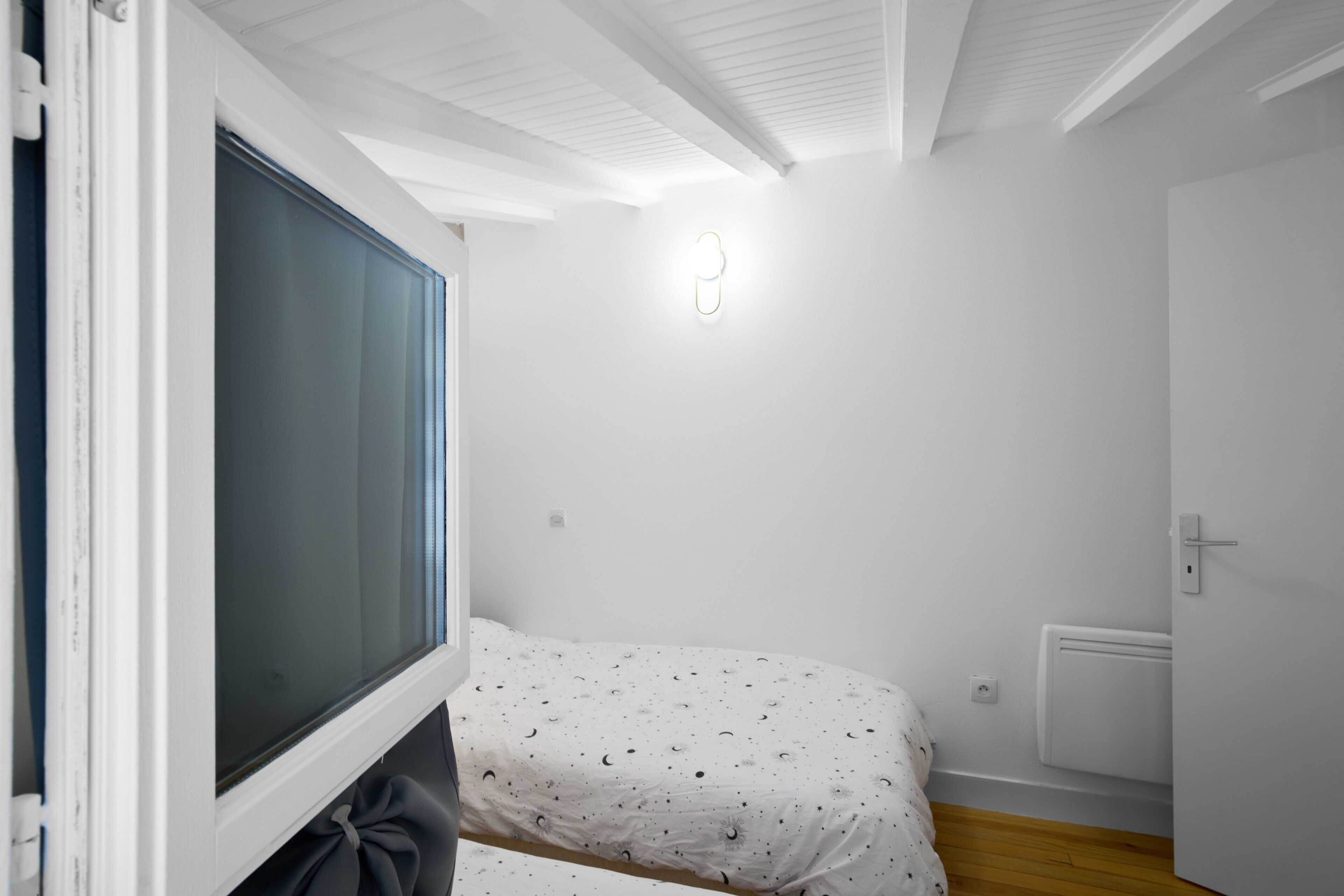 A bedroom with a bed, a window, a door, and a light fixture on the wall.