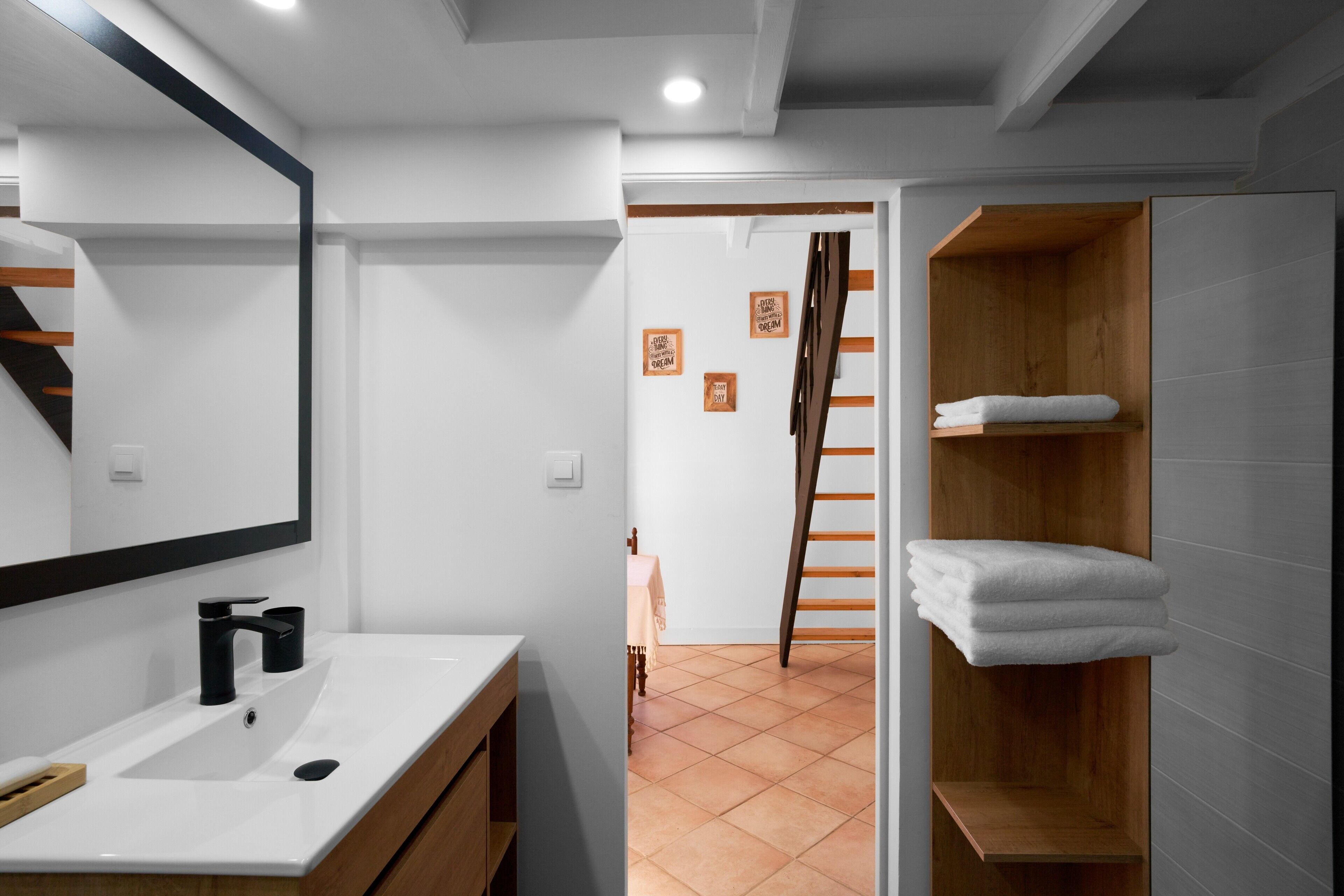 A modern bathroom with a white sink, mirror, and wooden vanity. A wooden ladder leads to a lofted sleeping area with a wooden shelf and a white towel.